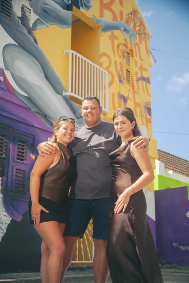 Two women and a man posing together in front of a colorful mural in Curacao