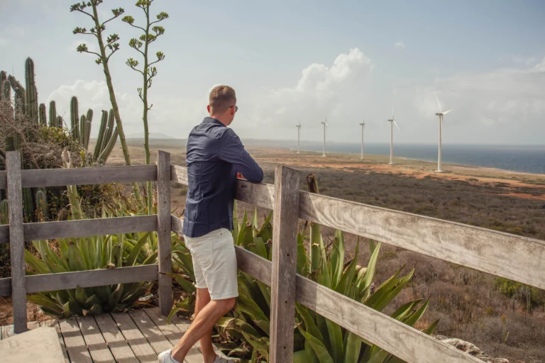 Man standing on a wooden boardwalk overlooking the Curacao coastline with wind turbines in the distance
